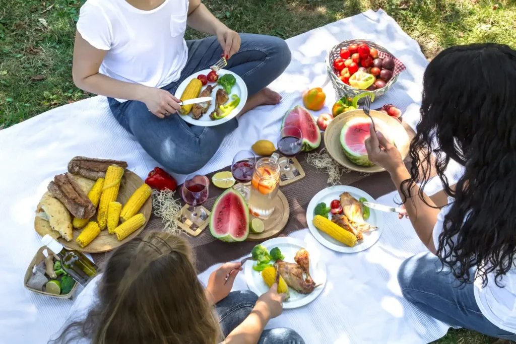 Healthy summer picnic with fresh fruits, whole-grain sandwiches, vegetable snacks, and bottled water arranged on a picnic blanket in a park.